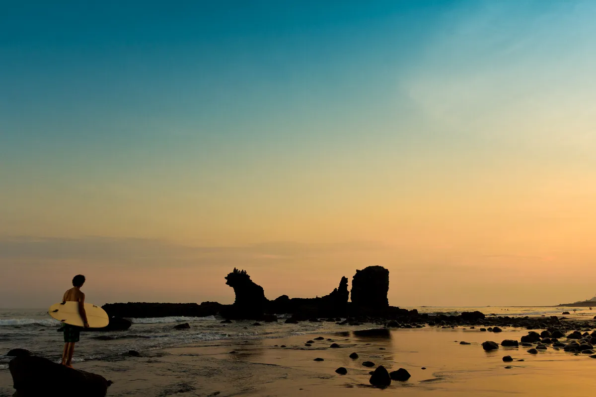 A surfer silhouetted by El Tunco rock at sunset, El Salvador